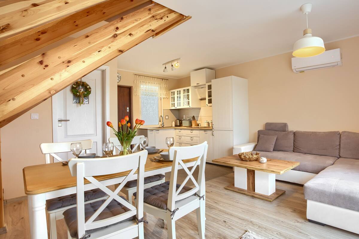 A modern living room with a wooden ceiling, a dining area with white chairs and a table, and a grey sofa.