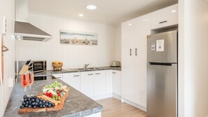 A modern kitchen with white cabinets, a stainless steel refrigerator, a fruit and cheese board, and a painting on the wall.