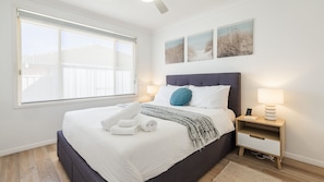 A neatly made bed with a headboard, a bedside table with a lamp, and a window with blinds.