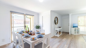 A dining area with a table set for six, a white console table with a mirror, and a television in the adjacent room. - Pet friendly Driftwood Cottage, 1-28 Farm Rd - W-Fi, Air Con and ground floor (Fingal Bay)