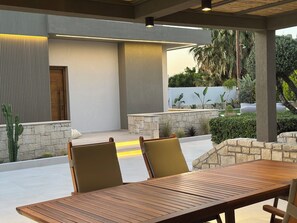 An outdoor patio with a wooden table and chairs, stone walls, and a view of greenery.