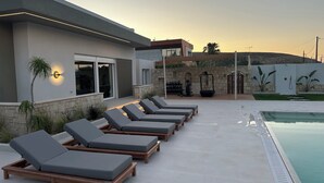 A poolside area with lounge chairs, a stone wall, and a building with a modern design. - Άνετη βίλα οικογενειακού μεγέθους με υδρομασάζ! (Heraklion, Crete)