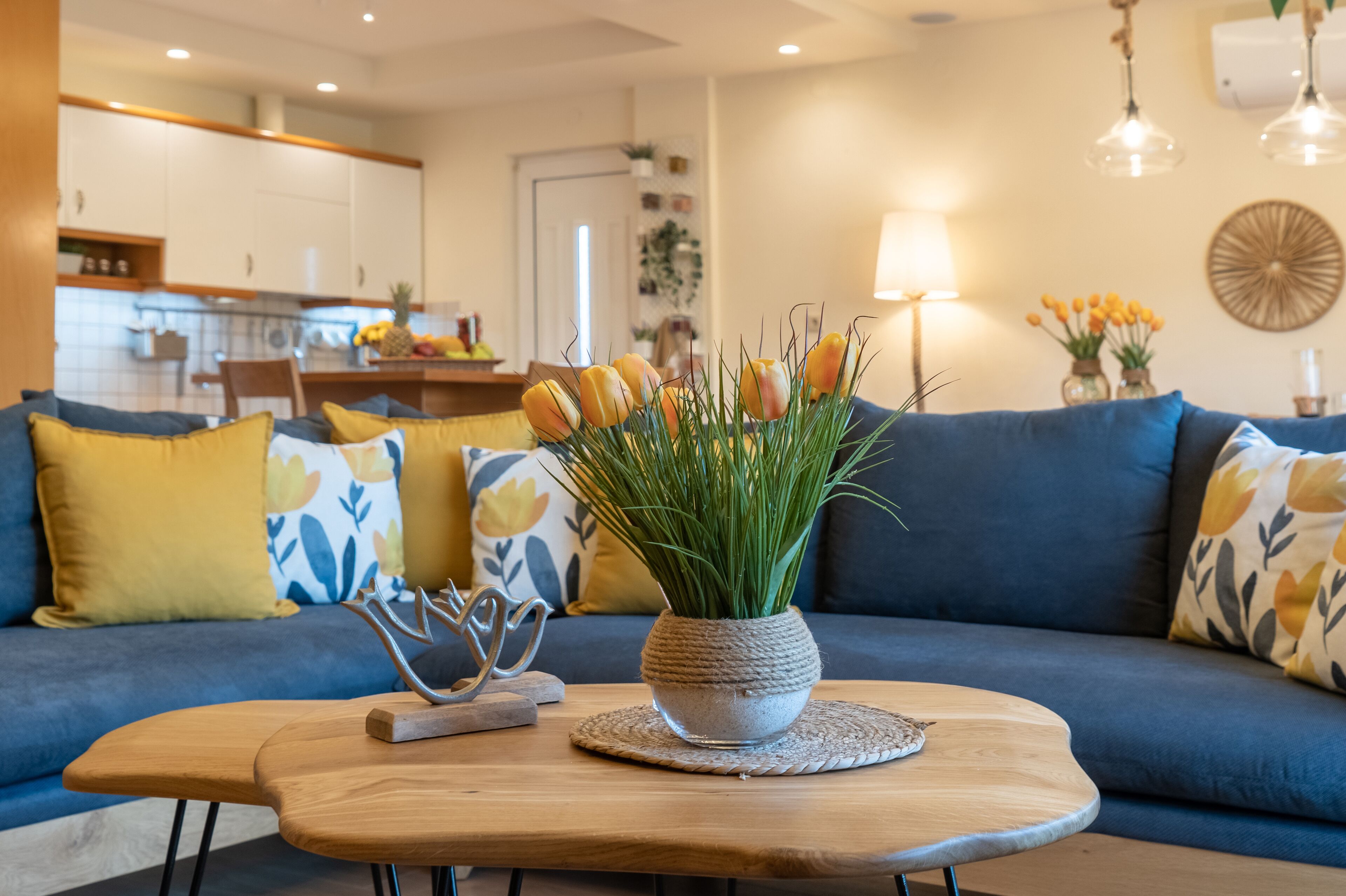 A living room with a blue sofa, yellow and white patterned pillows, a wooden coffee table with a potted plant, and a kitchen in the background.