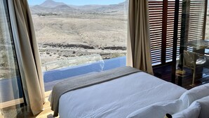 A bedroom with a large bed, a view of a desert landscape, and a balcony with a table and chair. - Wake up to nature in this modern glass house (fuerteventura)
