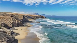 A coastal landscape with a sandy beach, rocky cliffs, and a clear blue sky. - Wake up to nature in this modern glass house (fuerteventura)