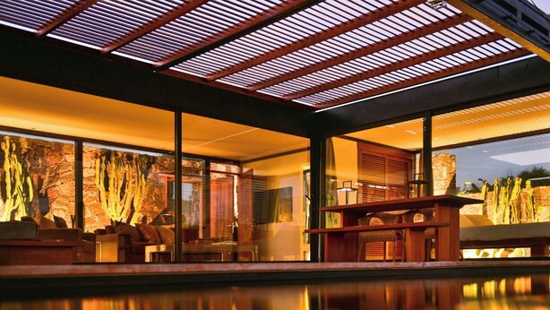 A modern indoor pool area with a glass enclosure, stone walls, and a seating area with chairs and a table. - Wake up to nature in this modern glass house (fuerteventura)