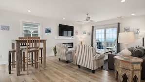 A modern living room with a wooden dining table, striped armchairs, a fireplace, and a view of the sea through large windows.