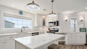 A modern kitchen with white cabinetry, a large island, pendant lighting, and a view of the outside through a window.