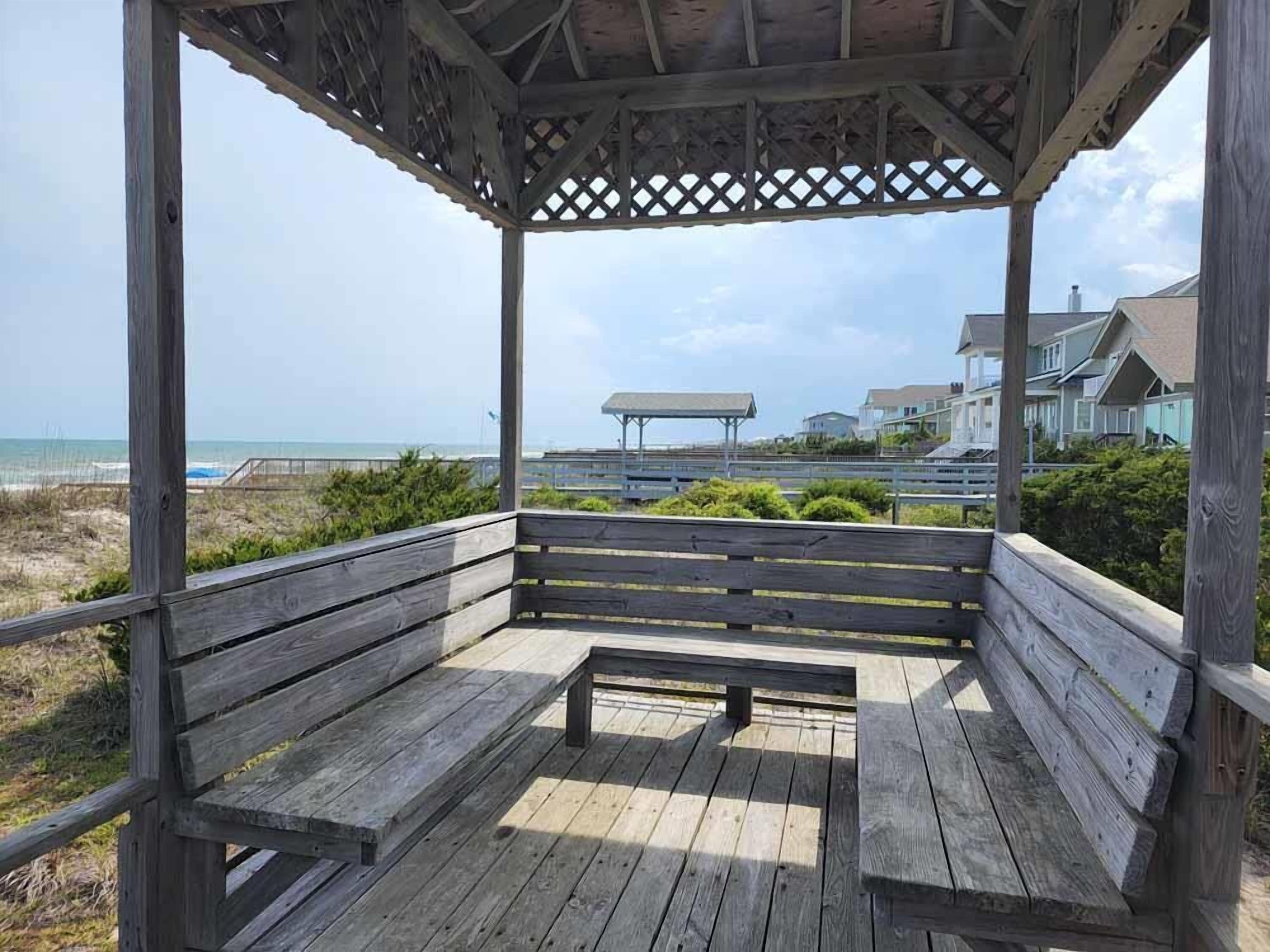A wooden gazebo with benches overlooking a beach and houses.