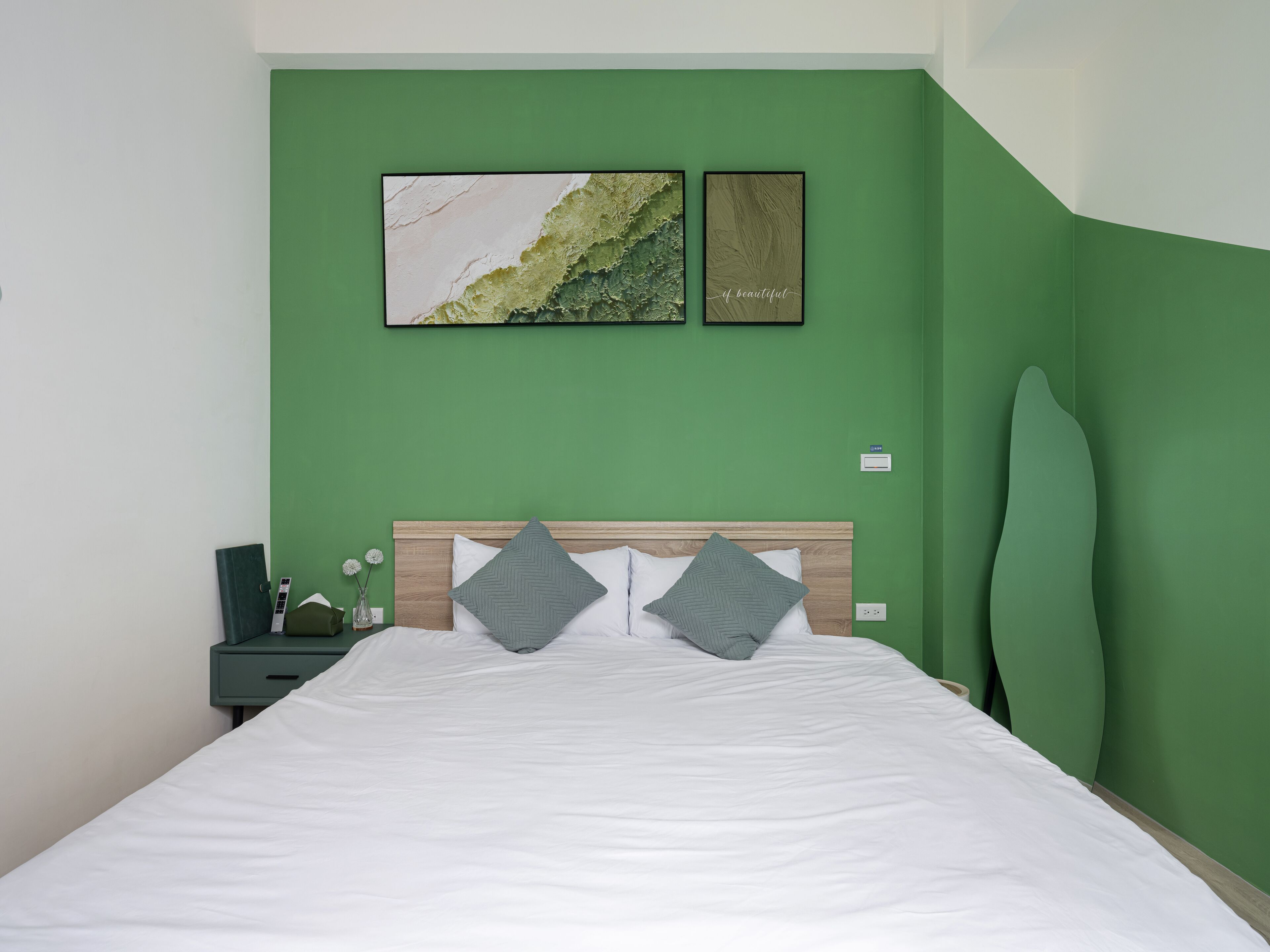 Un dormitorio con pared verde, cabecera de madera, cubrecama blanco y dos almohadas. Hay dos cuadros enmarcados en la pared.