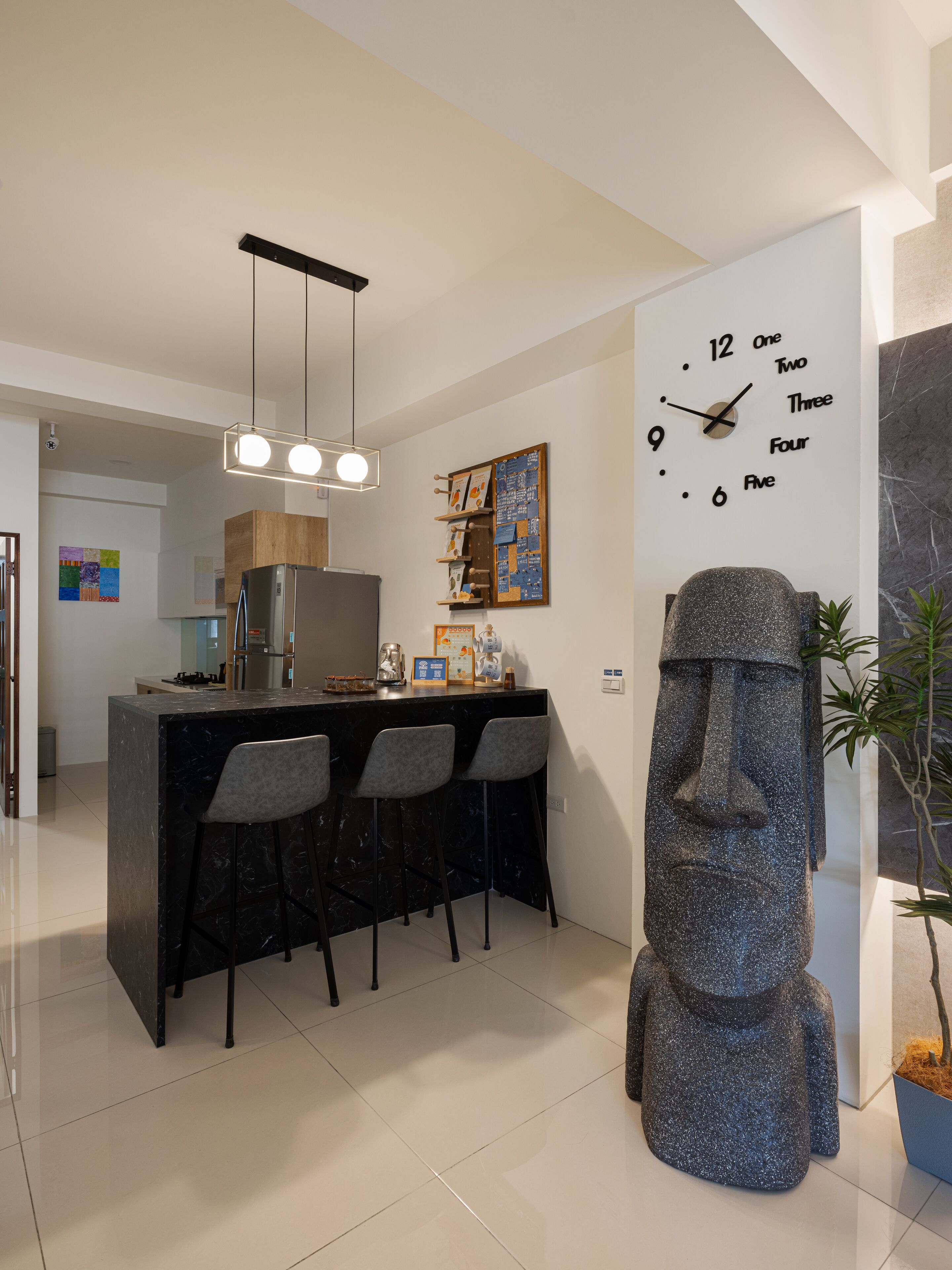 A modern kitchen with a bar area, a large clock on the wall, and a statue of a moai.