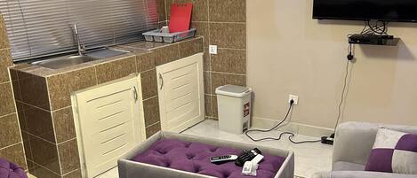A modern living room with a kitchen area, featuring a sofa, ottoman, and a flat-screen TV mounted on the wall.