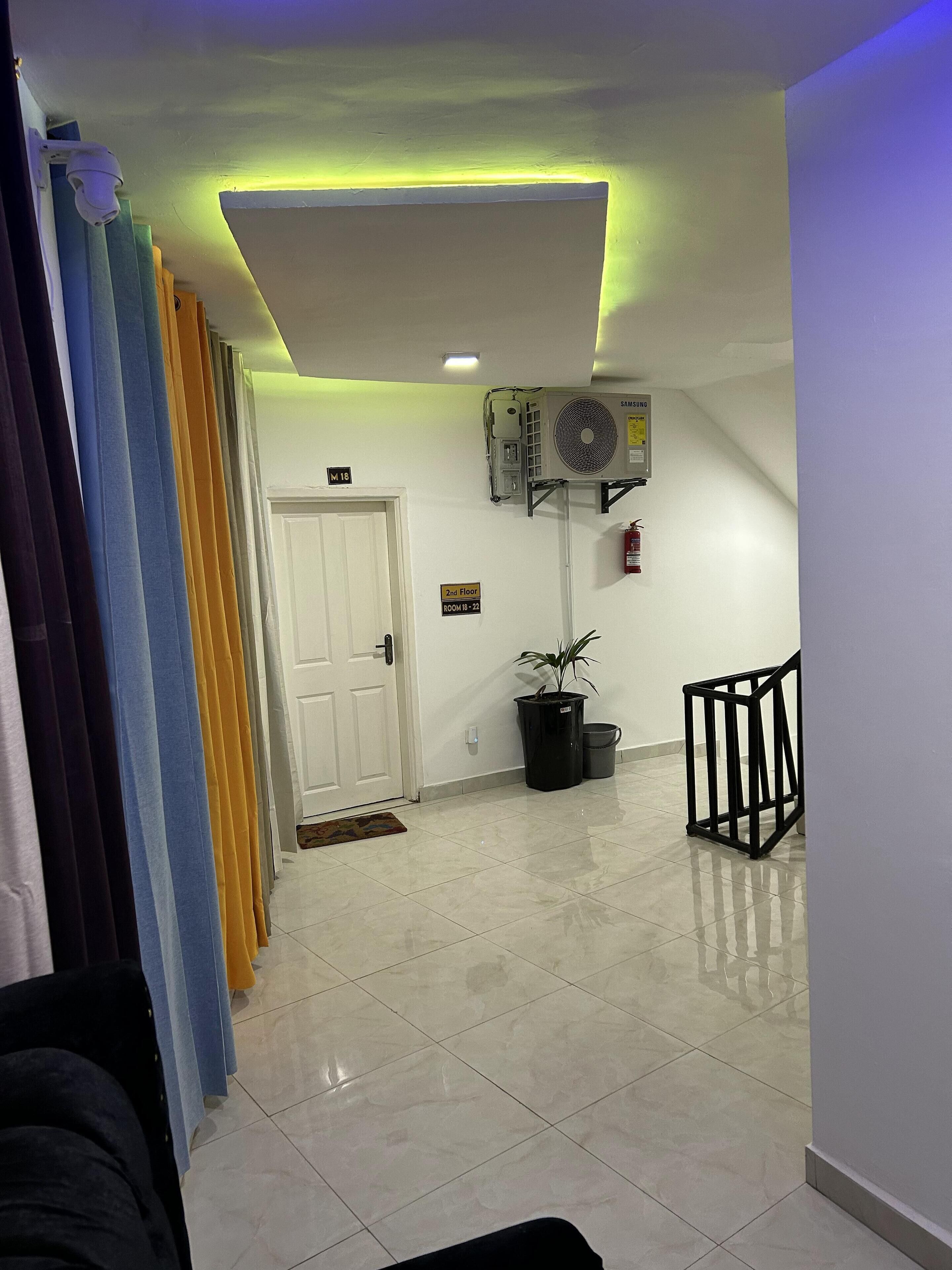A hallway with a white door marked "Room 18," a sign indicating "2nd Floor Room 18-22," a Samsung air conditioner, a potted plant, a trash bin, and a black metal chair.
