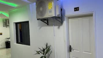 Un passadís amb una porta blanca, un aparell d'aire condicionat fixat a la paret, una planta en test, un cubell de brossa i una catifa de benvinguda.