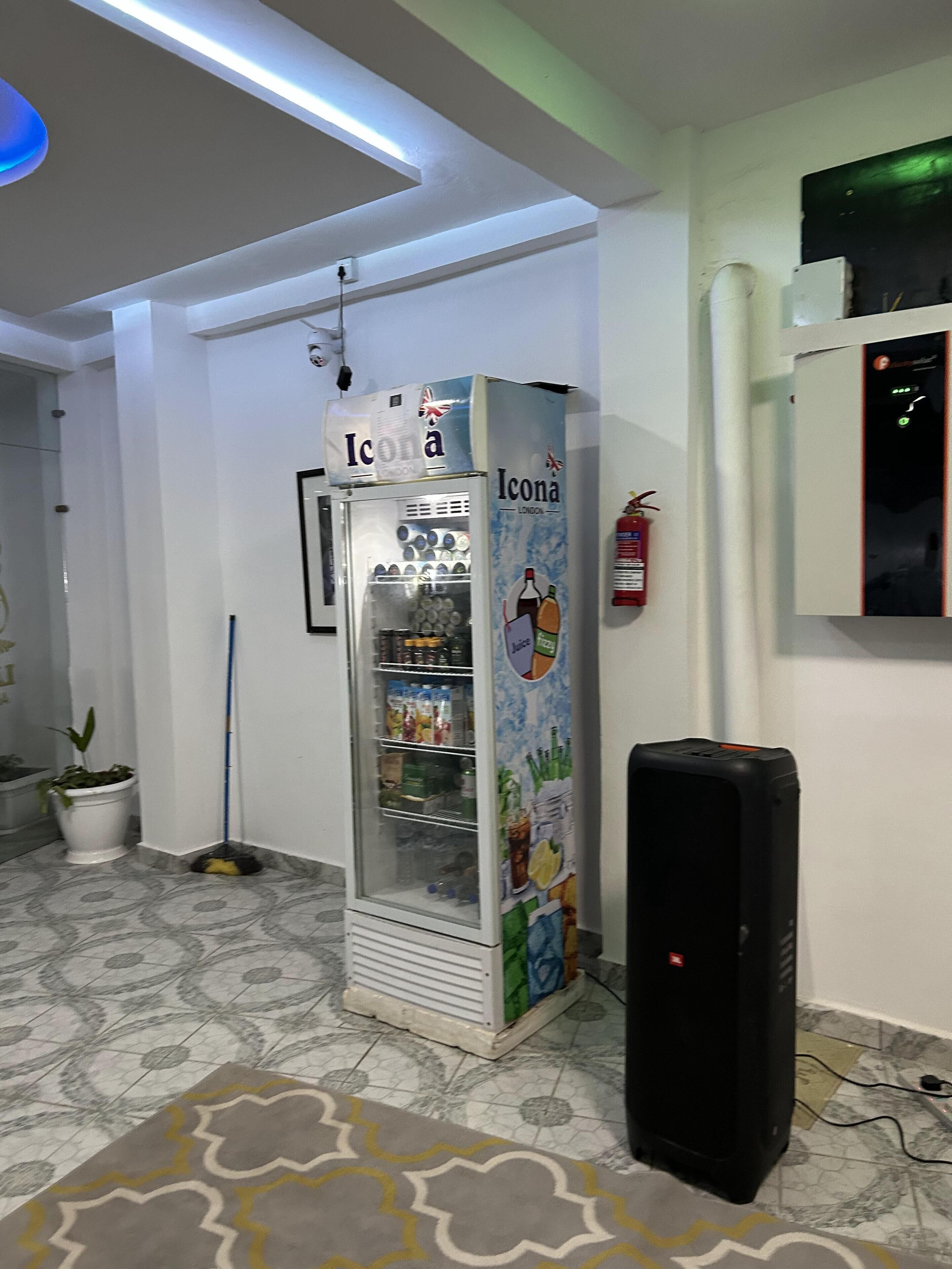 A room with a refrigerator stocked with beverages, a mop, a potted plant, and a speaker.