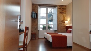 A hotel room with a wooden floor, a bed with a red blanket, a TV mounted on the wall, and a stone wall. - Pensión O Códice (Santiago de Compostela)