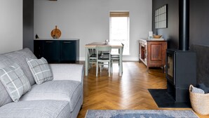 A modern living room with a grey sofa, wooden flooring, a dining area with a table and chairs, a kitchen with dark cabinets, and a black fireplace. - Meadow View Cottage (Carnforth)