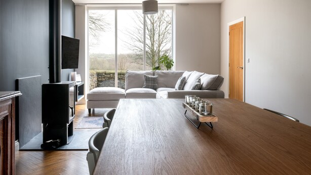 A modern living room with a wooden dining table, a grey sofa, and a TV mounted on the wall. - Meadow View Cottage (Carnforth)