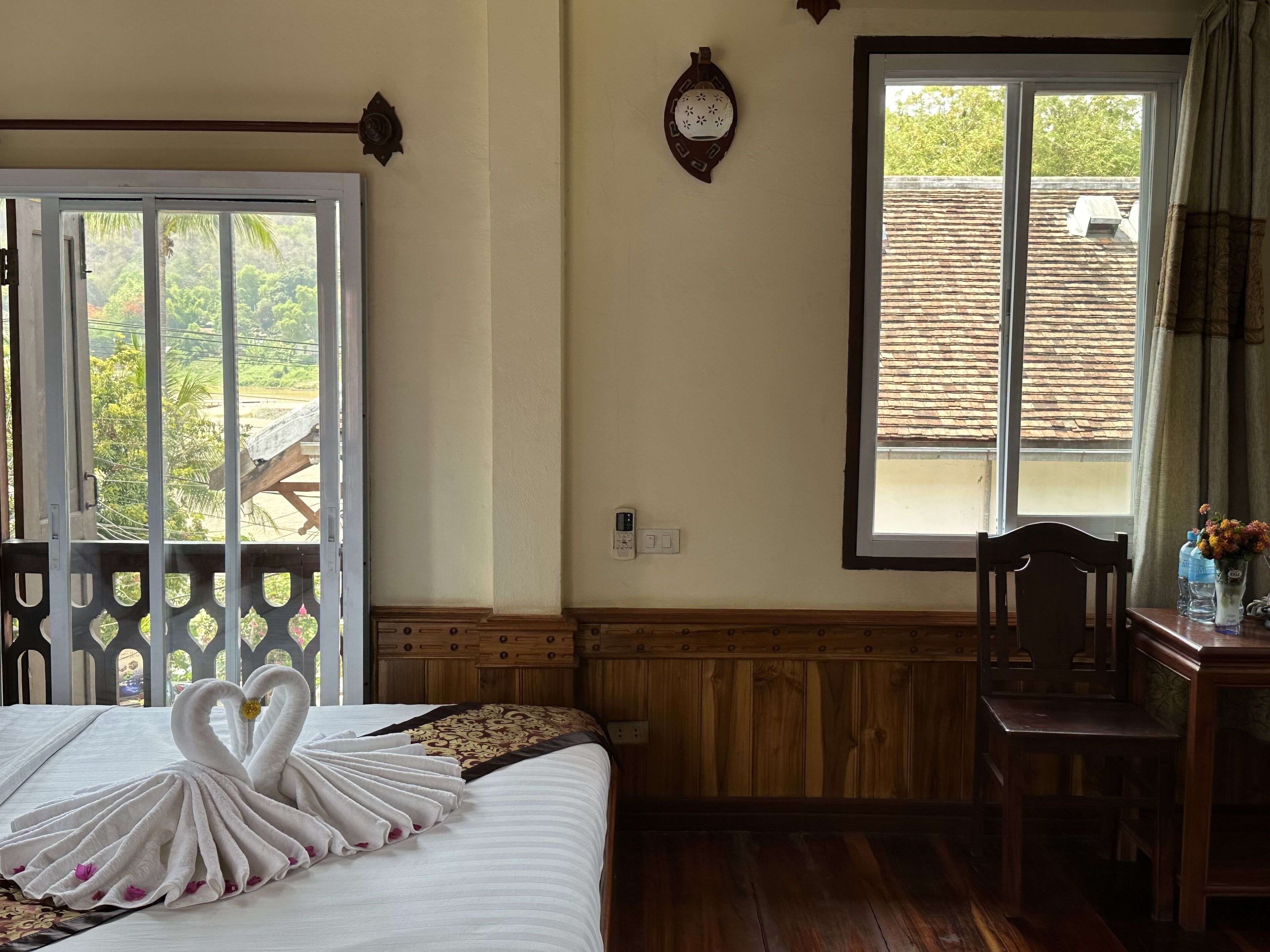 A hotel room with a bed, a wooden chair, a table with water bottles, a window with a view of trees and a building, and a wall clock.