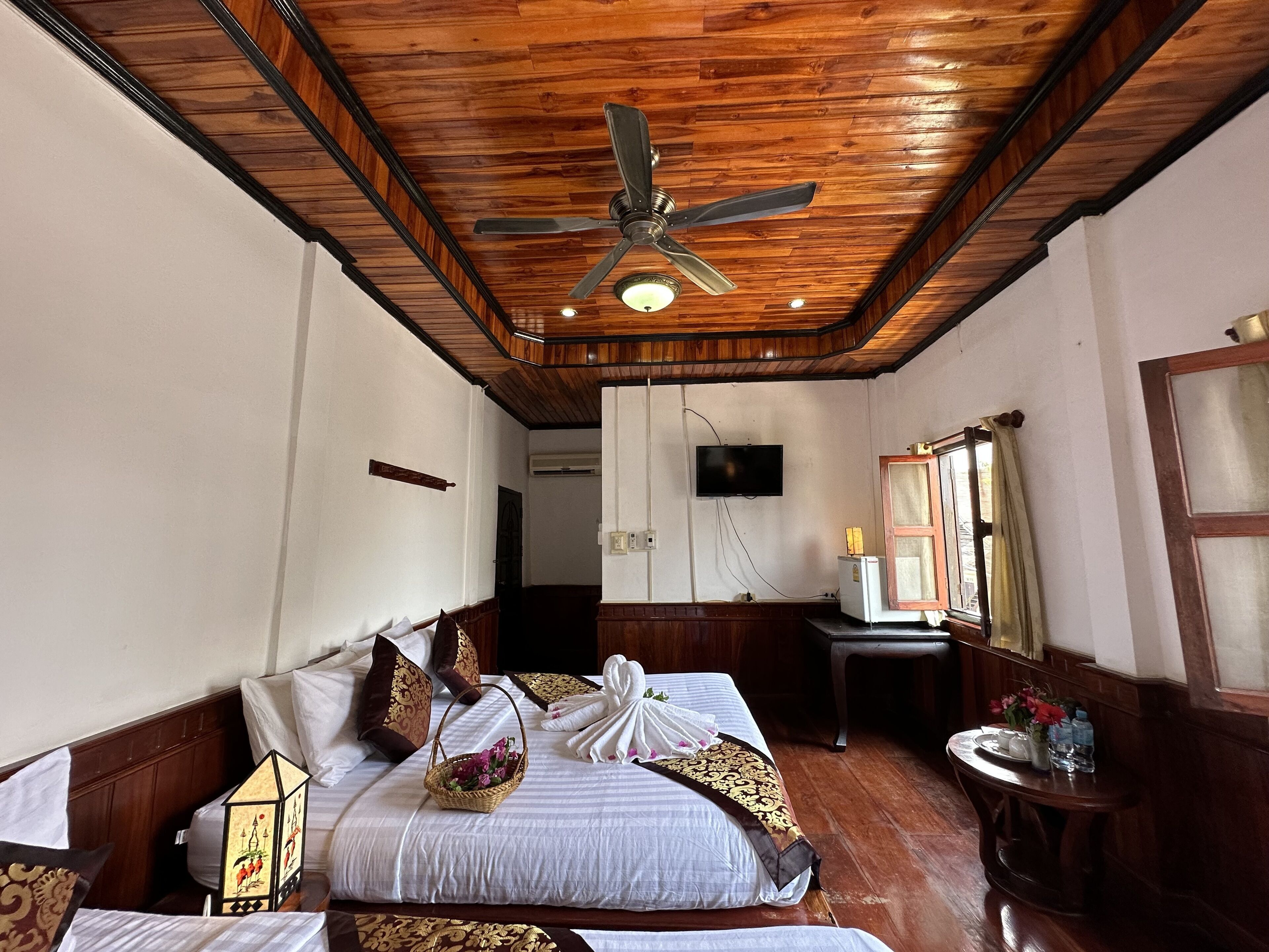 A hotel room with a wooden ceiling, a bed with white bedding and decorative pillows, a small table with a vase of flowers, and a television mounted on the wall.
