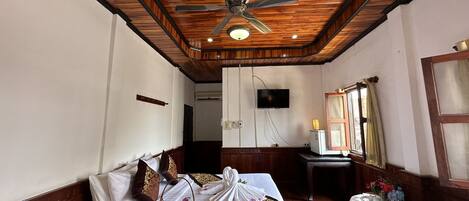 A hotel room with a wooden ceiling, a bed with white bedding and decorative pillows, a small table with a vase of flowers, and a television mounted on the wall.