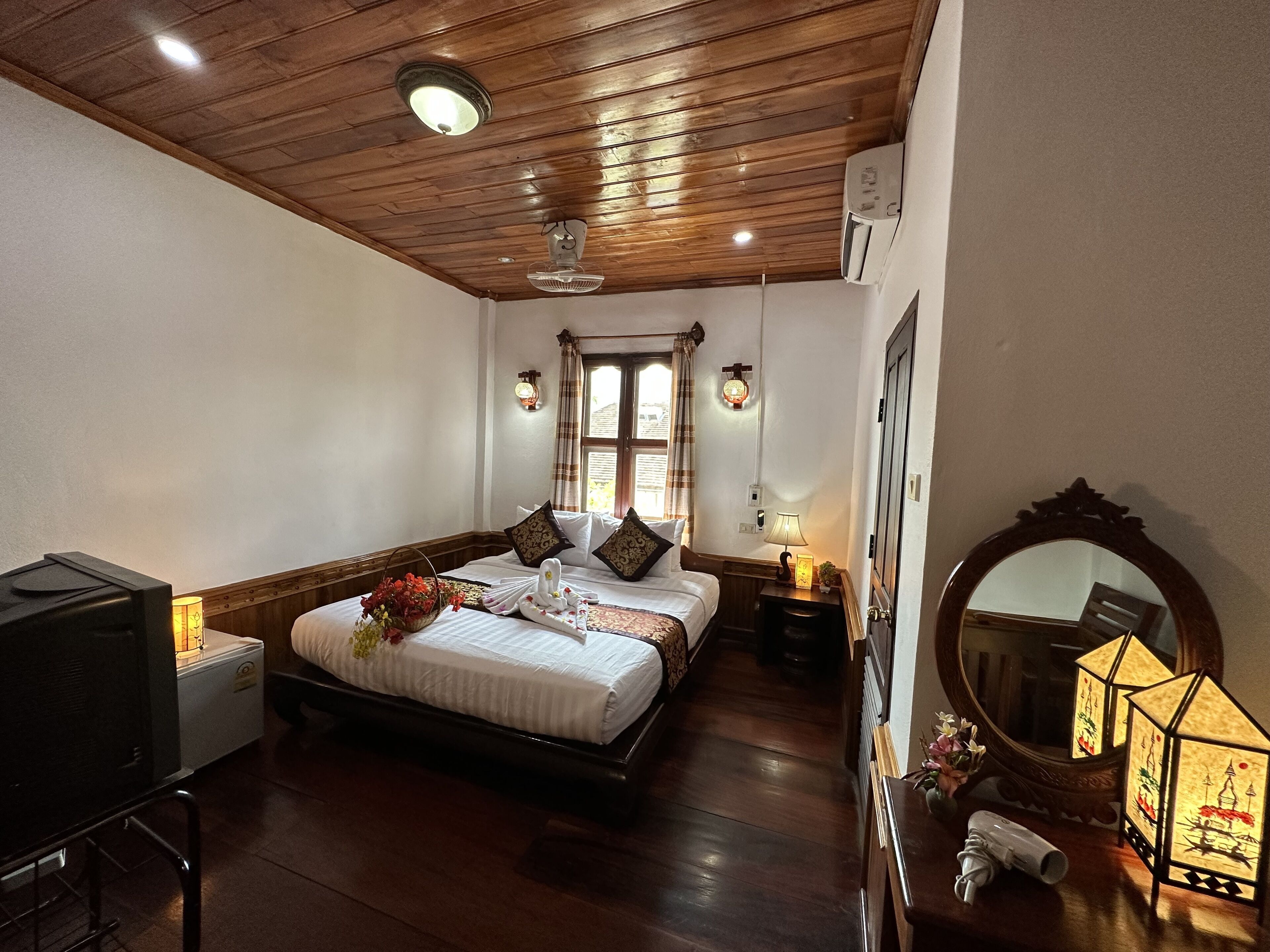 A bedroom with a wooden ceiling, a bed with white linens and decorative pillows, a bedside table with a lamp, a mirror, and a small table with a decorative item.