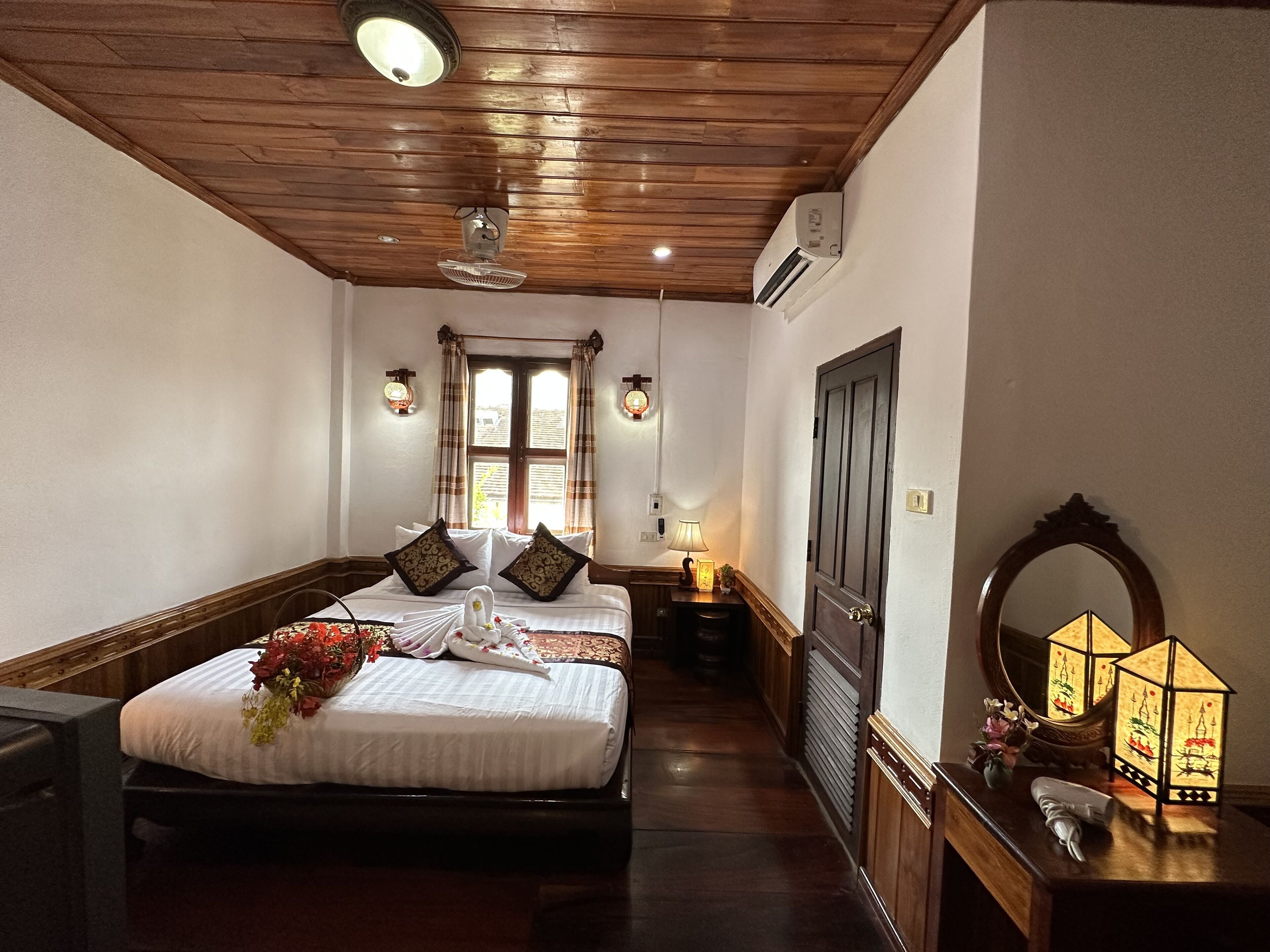A hotel room with a wooden ceiling, a large bed with white bedding and decorative pillows, a bedside table with a lamp, a mirror, and a decorative lantern.