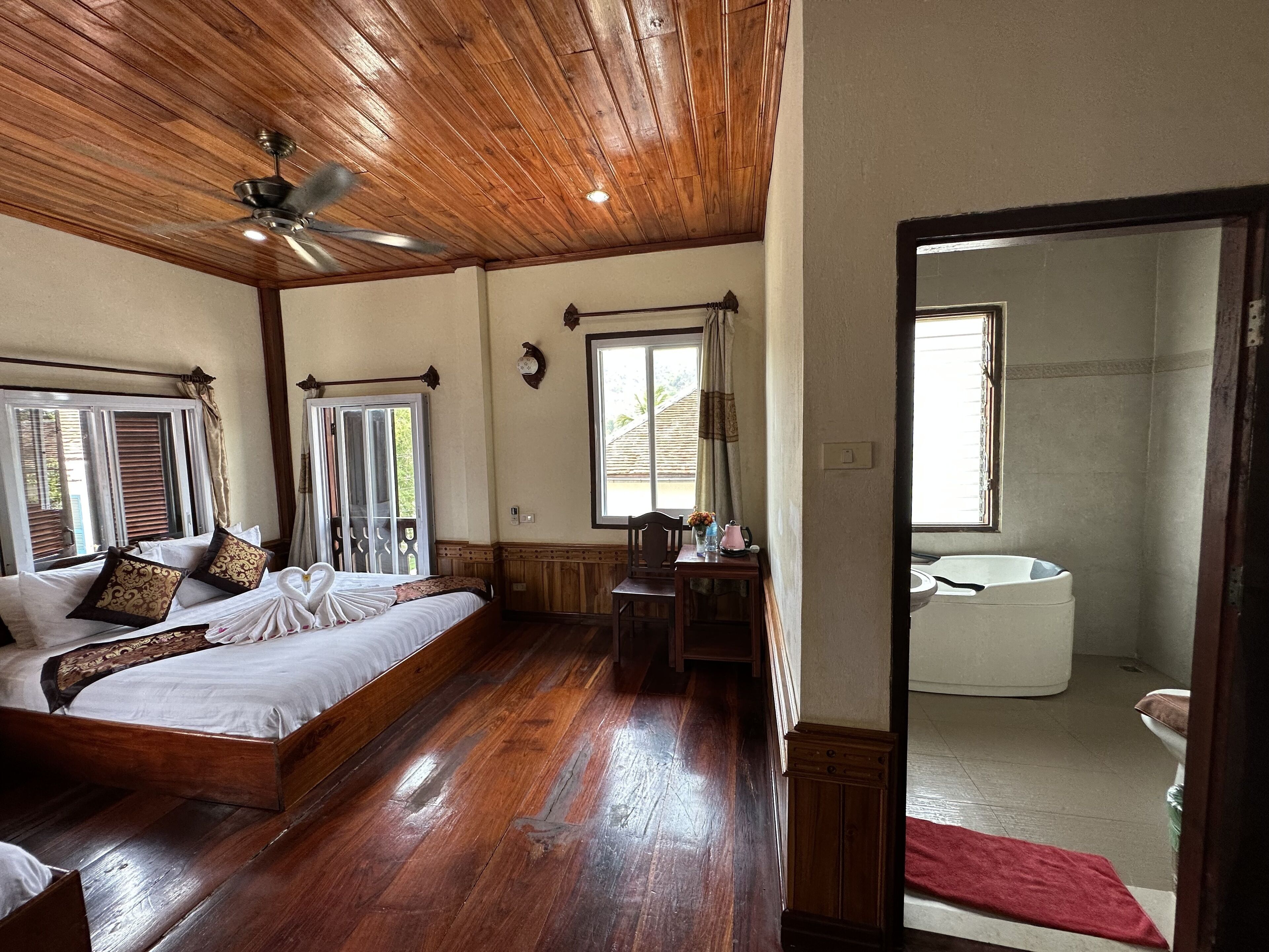 A room with a wooden ceiling, a bed with white bedding, a wooden floor, a small table with a vase, and a window with curtains.