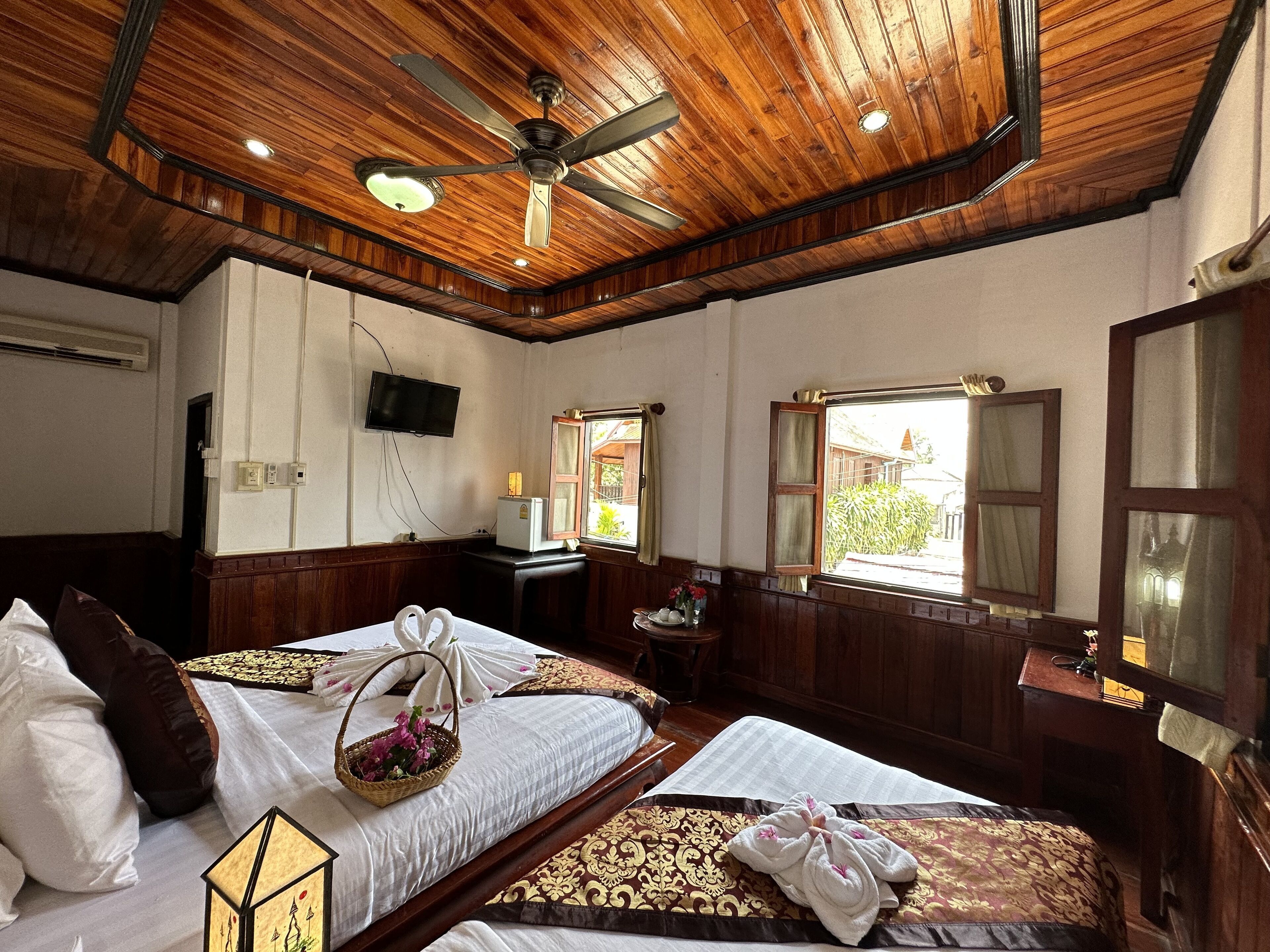 A hotel room with two beds, a ceiling fan, a TV, a window, and a small desk with a lamp.