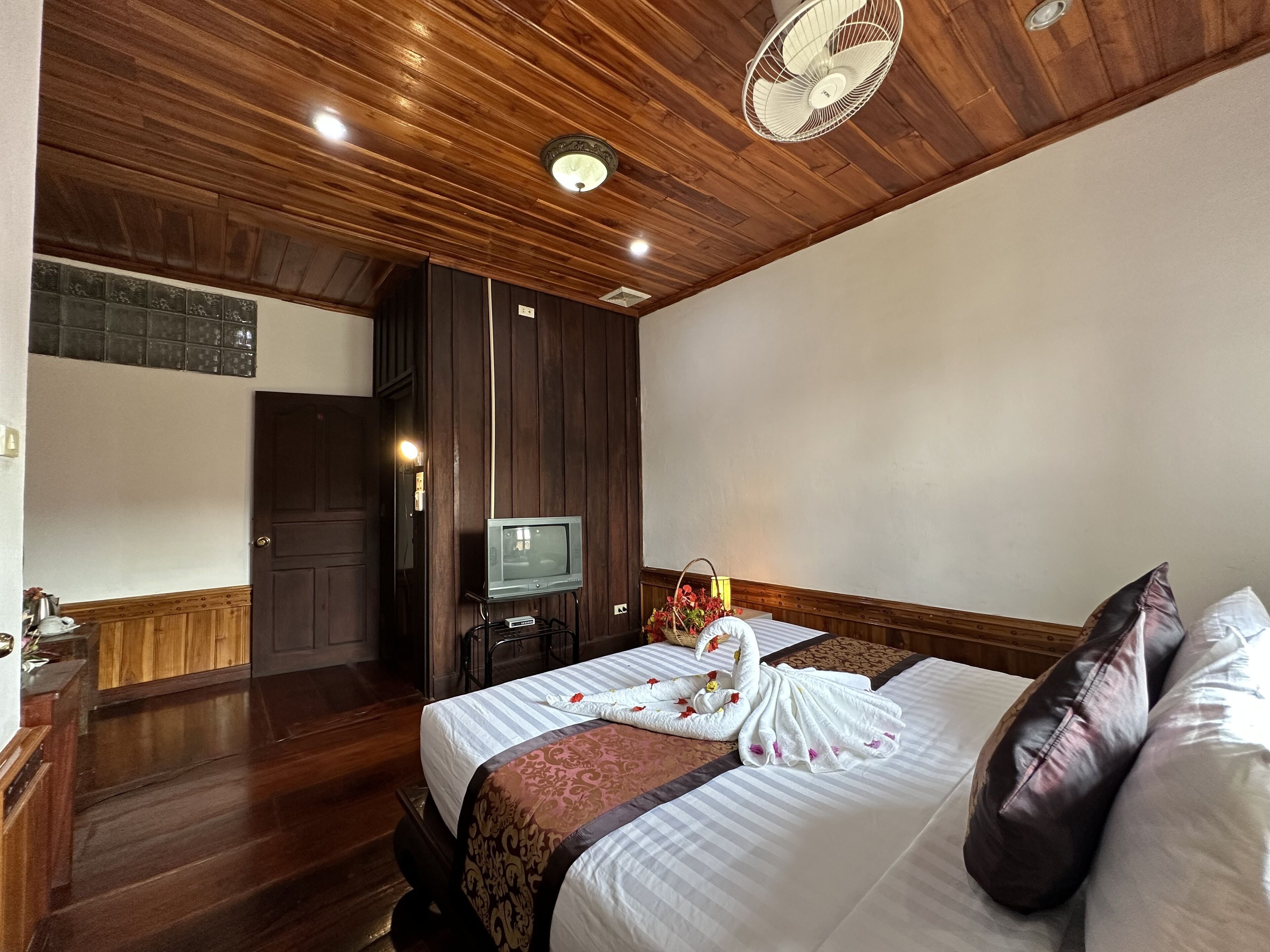 Een hotelkamer met een houten plafond, een bed met een wit-bruine spreideken, een televisie op een standaard en een ventilator.