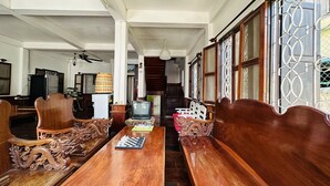 A traditional wooden dining table with chairs, a television, and a fan in a spacious room with large windows and a staircase in the background. - Villa Thida Mekong Riverside (Luang Prabang)