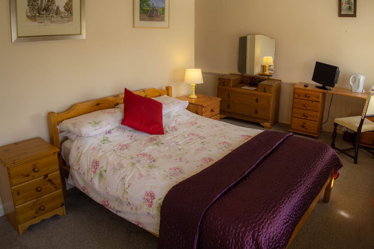 A bedroom with a bed, wooden nightstands, a dresser, a desk with a computer, and a lamp.