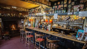 A bar with wooden stools, a fireplace, and shelves stocked with bottles and glasses. - Duke of York (Winkleigh)