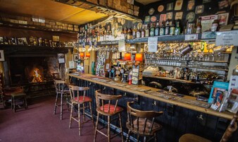Um bar com bancos de madeira, lareira e prateleiras repletas de garrafas e copos.