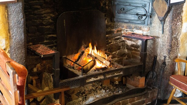 A stone fireplace with a fire burning, a wooden bench, and a rustic wooden table with a metal tray. - Duke of York (Winkleigh)
