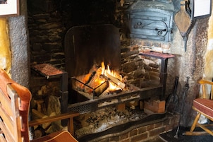 Lareira de pedra com fogo aceso, um banco de madeira e uma mesa rústica de madeira com uma bandeja de metal.