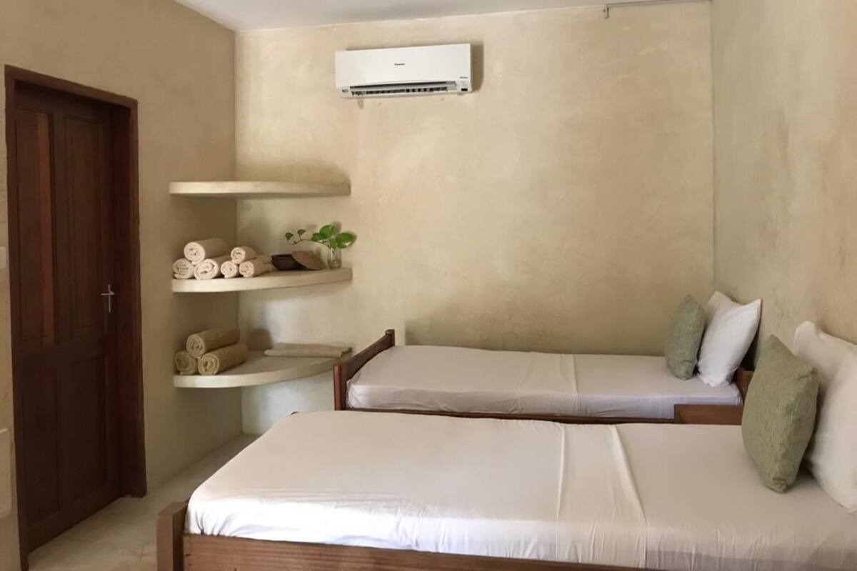 A room with two beds, a wooden door, a shelf with towels, and an air conditioning unit.