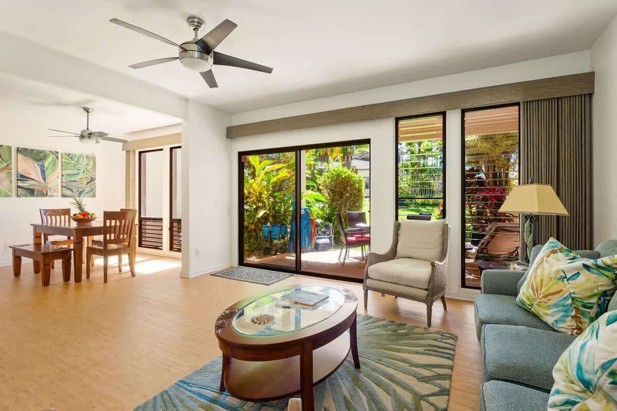 A spacious living area with a dining table, a sofa, and a coffee table. There is a ceiling fan, a large window with a view of greenery, and a door leading to an outdoor area.