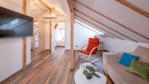 A modern loft bedroom with a sloped ceiling, wooden beams, a flat-screen TV, a sofa, a red chair, and a small table with a plant. - Bohemian Guest House (Sofia)