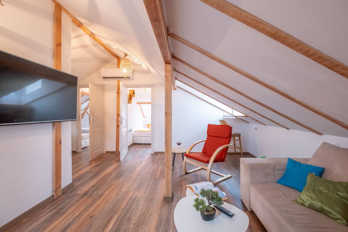 A modern loft bedroom with a sloped ceiling, wooden beams, a flat-screen TV, a sofa, a red chair, and a small table with a plant.