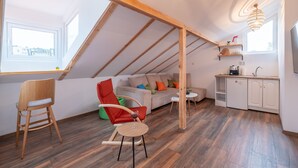 A cozy attic room with a wooden ceiling, a sofa, a dining table, a kitchen area, and a window providing natural light. - Bohemian Guest House (Sofia)