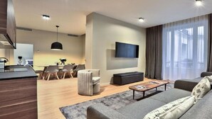 Ein modernes Wohnzimmer mit einer Couch, einem Couchtisch und einem an der Wand montierten Fernseher. Es gibt einen Essbereich mit Tisch und Stühlen sowie eine Küchenzeile mit Arbeitsfläche und Geräten.