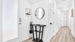A hallway with white walls, a wooden floor, a round mirror, a black console table, and a potted plant.