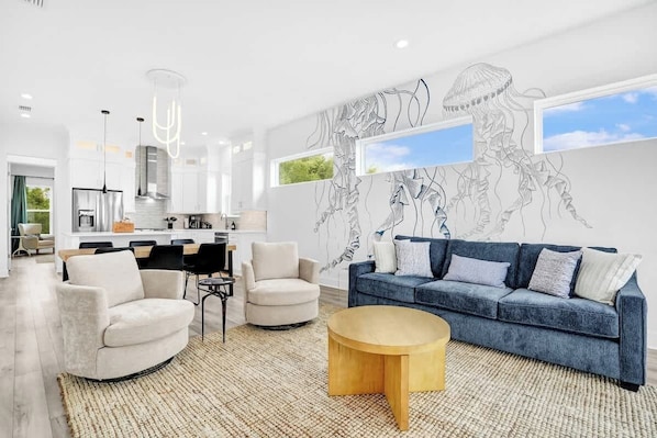 A modern living room with a blue sofa, two armchairs, a wooden coffee table, and a kitchen in the background.