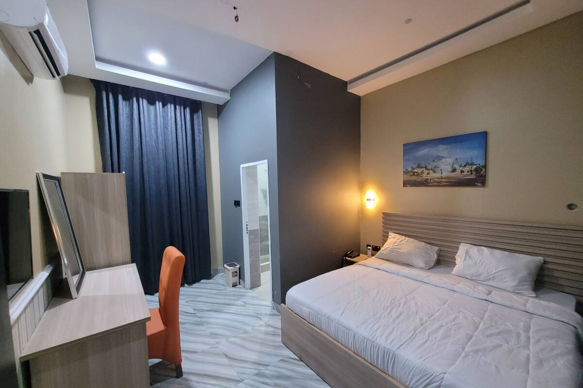 A hotel room with a bed, a desk, an orange chair, a painting on the wall, and a door leading to another room.