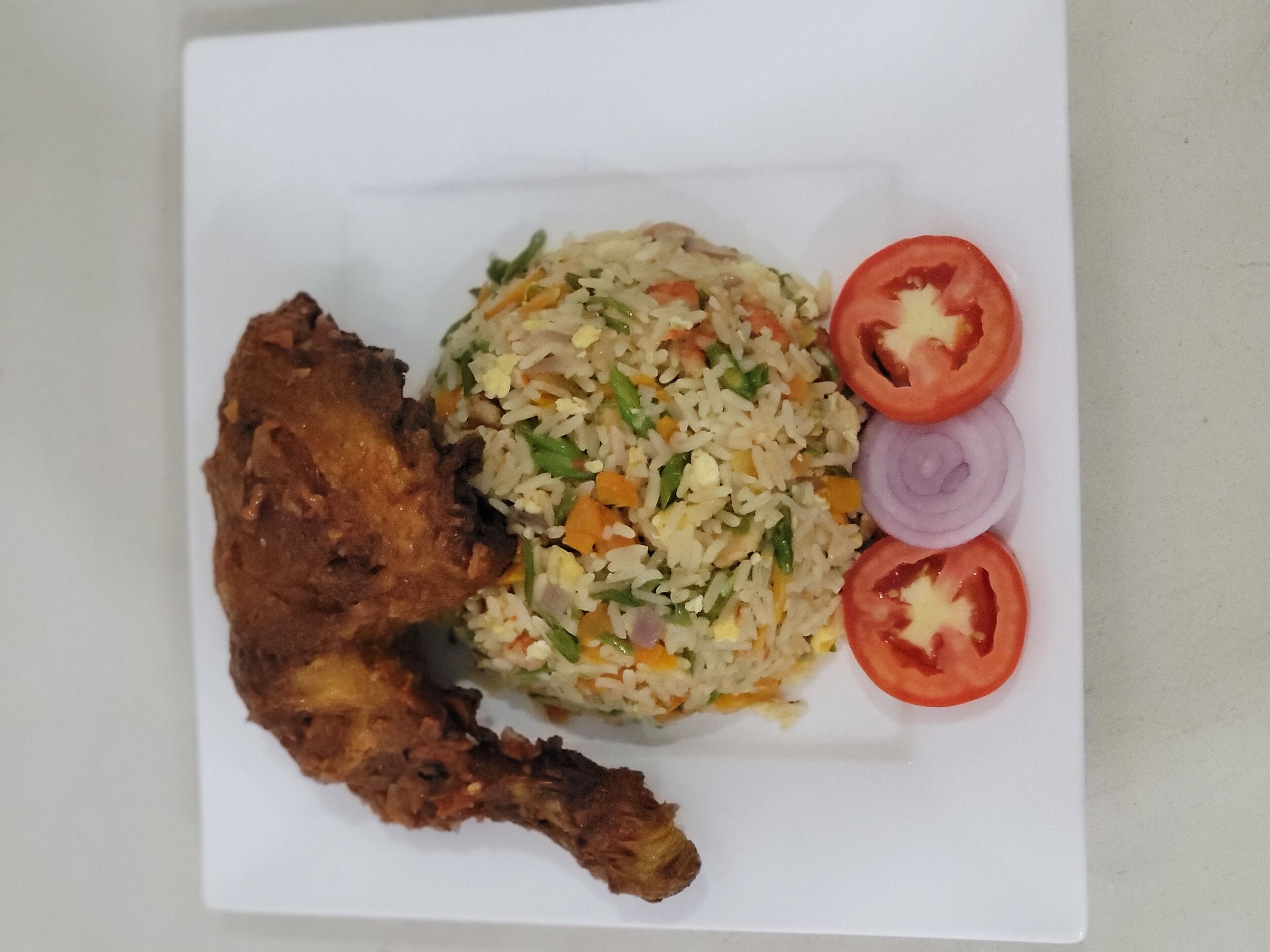 A plate of fried chicken, fried rice with vegetables, and sliced tomatoes and onions.