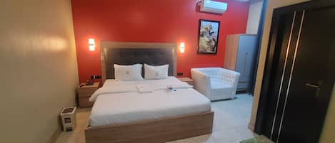 Una habitación de hotel con una cama grande, una mesita de noche, un sillón blanco, un armario y una puerta.