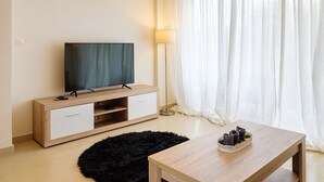A modern living room with a flat-screen TV on a wooden TV stand, a floor lamp, a black furry rug, and a wooden coffee table with a decorative bowl. - Marathon Luxury Suites - Schinias Beach (Marathon)