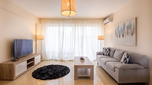 A modern living room with a flat-screen TV, a sofa, a coffee table, and a large window with sheer curtains. - Marathon Luxury Suites - Schinias Beach (Marathon)