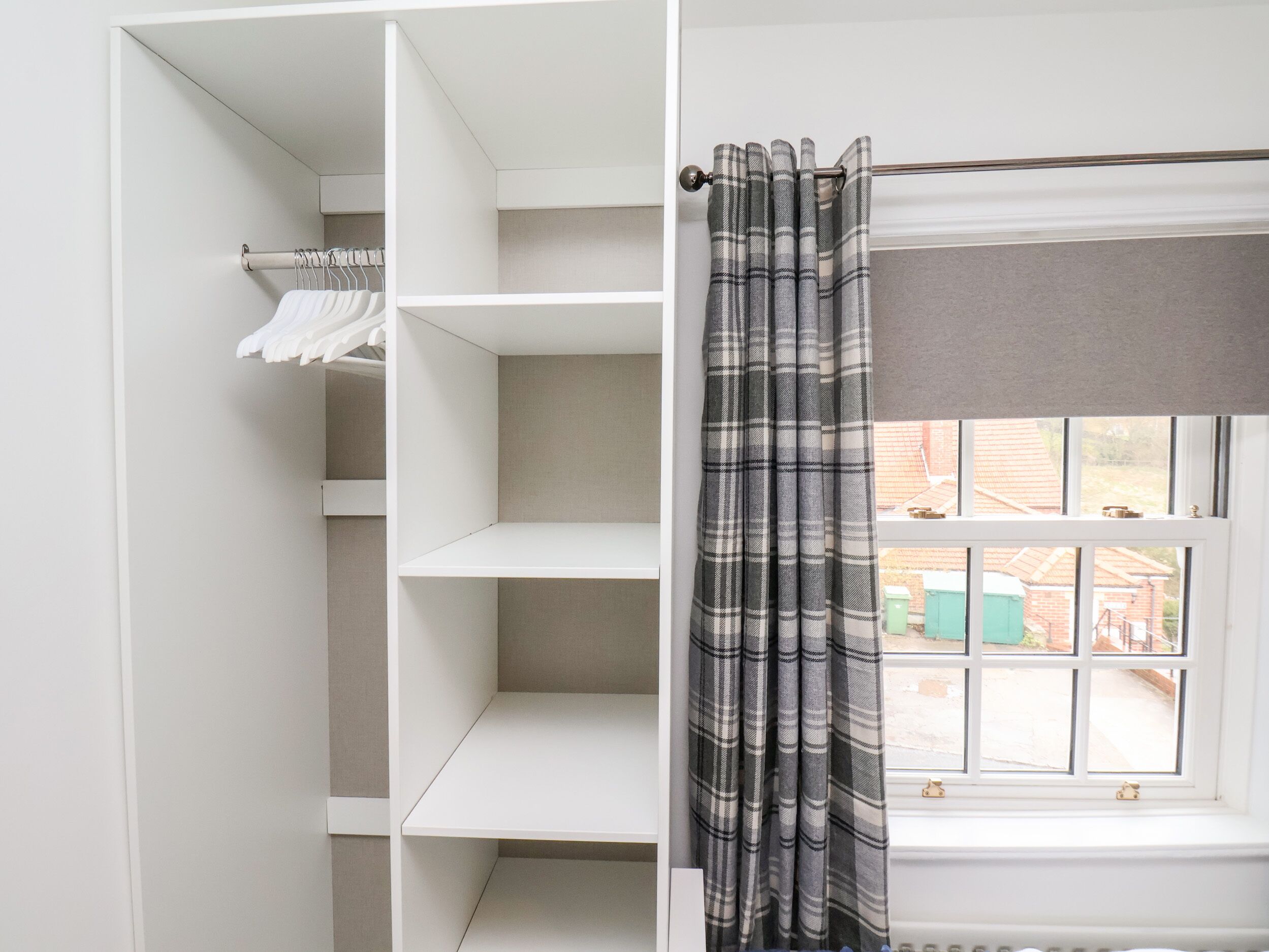 A room with a white wardrobe, a window with a view of the outside, and a plaid curtain.